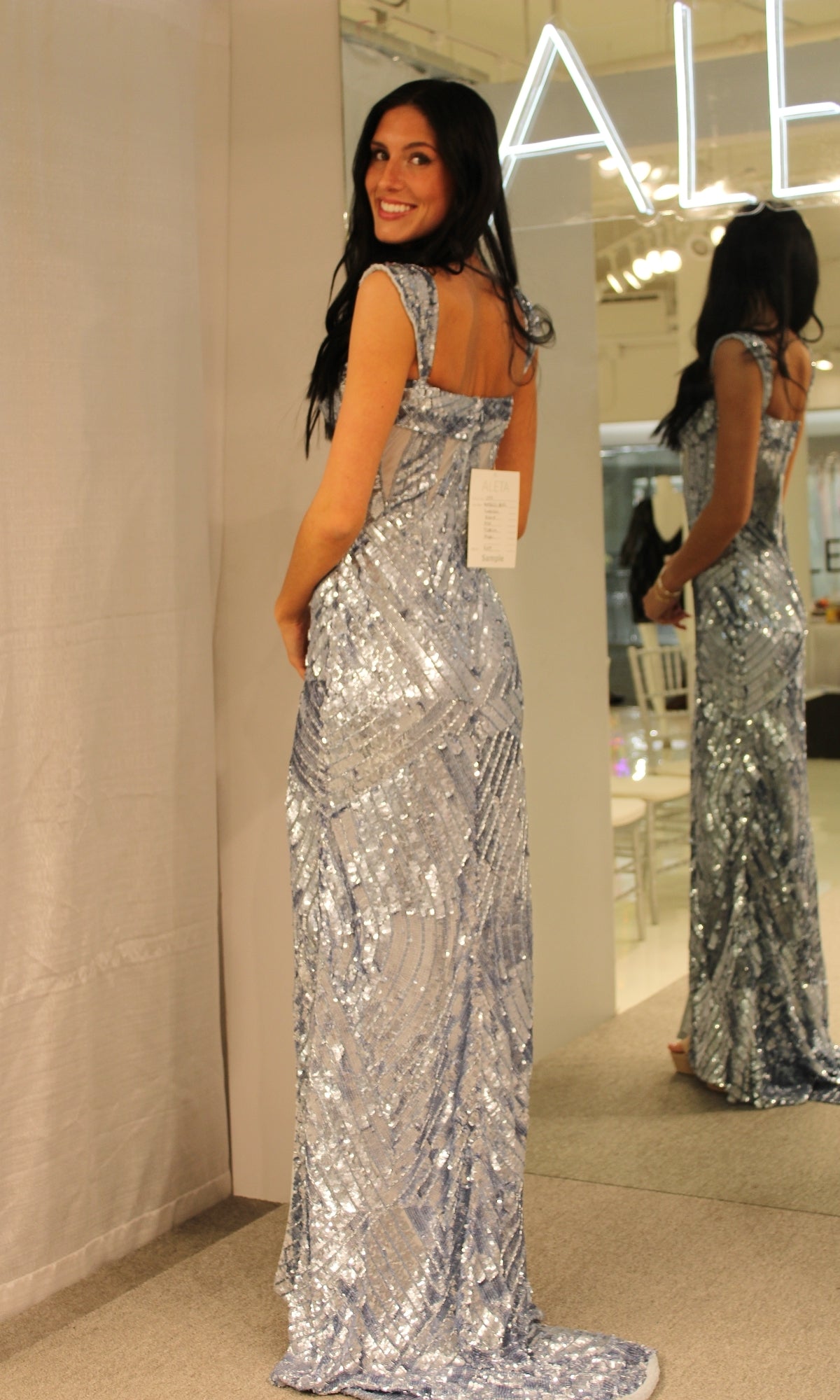 A woman stands in front of a fitting room mirror, smiling over her shoulder in the Cap-Sleeve Long Sequin Prom Dress 1173—a floor-length silver gown with cap sleeves and a tag still attached.